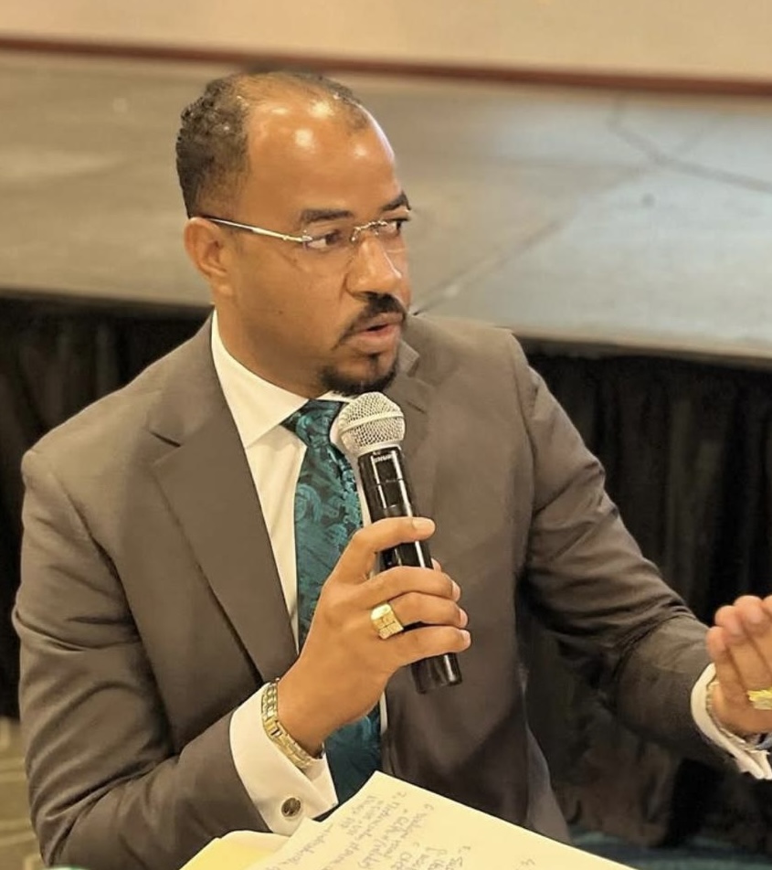 Un homme en costume gris tenant un micro, parlant lors d'un événement, avec des notes sur une table devant lui.