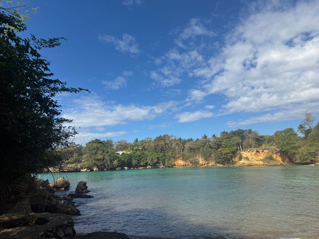 Vue d'une plage tranquille avec une eau turquoise, des rochers et une végétation luxuriante sous un ciel bleu avec quelques nuages.